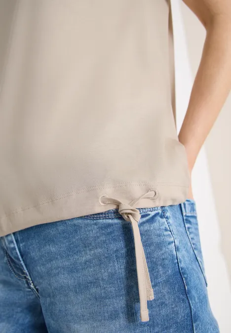 Nahaufnahme einer Person in beigem Oberteil mit Tunnelzug am Saum und blauen Jeans, Hand in Tasche.